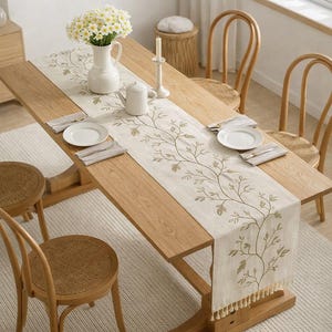 Può includere: Un tavolo da pranzo in legno chiaro con una tovaglia color crema con un motivo a foglie. Piatti bianchi, tovaglioli e posate sono apparecchiati per il pasto. Un vaso bianco con fiori e una teiera sono sul tavolo. Sedie in legno circondano il tavolo.