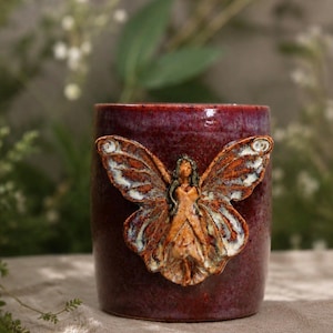 May include: A burgundy ceramic mug with a sculpted fairy design. The fairy has outstretched wings with brown and white details. The mug is cylindrical and has a glossy finish, ideal for holding beverages.