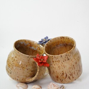 May include: Two handmade ceramic mugs with a speckled tan glaze. One mug has a handle with a red flower accent, and the other has a blue flower accent. Seashells are scattered at the base of the mugs.