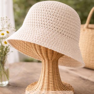May include: A cream-colored crochet bucket hat displayed on a wicker stand. The hat has a wide brim and is labeled "Crochet Bucket Hat Pattern Beginner Friendly" and "Boho Summer Hat Beginner Friendly". The background includes a vase of flowers and a woven bag.