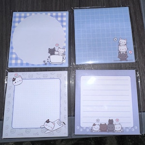 May include: Four square notepads with cat illustrations. The notepads have different designs, including a blue and white checkered pattern, a grid pattern, a dotted pattern, and lined paper. Each notepad features cute cat drawings in various poses.