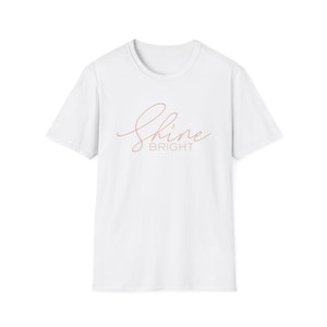 May include: A white t-shirt with a crew neck and short sleeves. The words "Shine Bright" are printed on the front in a light brown, cursive font. The word "Bright" is in a smaller, block font.