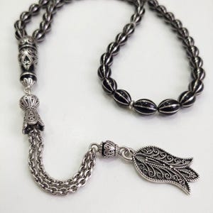 May include: An ornate silver-toned prayer bead necklace with black and silver beads. The necklace features detailed metalwork on the end pieces and a decorative pendant. The beads are oval-shaped and strung together with a chain.