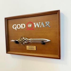 May include: A framed display featuring a replica of the "Blade of Olympus" from the "God of War" video game. The sword is mounted on a wooden plaque with the game's logo in silver and red. The blade has intricate gold and silver detailing.