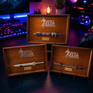 Op de afbeelding: Drie ingelijste decoratieve zwaarden uit The Legend of Zelda: Breath of the Wild. De houten frames tonen de zwaarden op zwarte standaards. De zwaarden zijn gelabeld met "Master Sword", "Edge of Duality" en "Windcleaver".