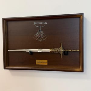 Może przedstawiać: Oprawiona ekspozycja repliki miecza. Miecz ma białe ostrze i zdobioną złotą rękojeść. Ekspozycja zawiera tekst "Elden Ring" i "Leda's Sword" na tle ciemnego drewna, zamknięta w brązowej ramie.