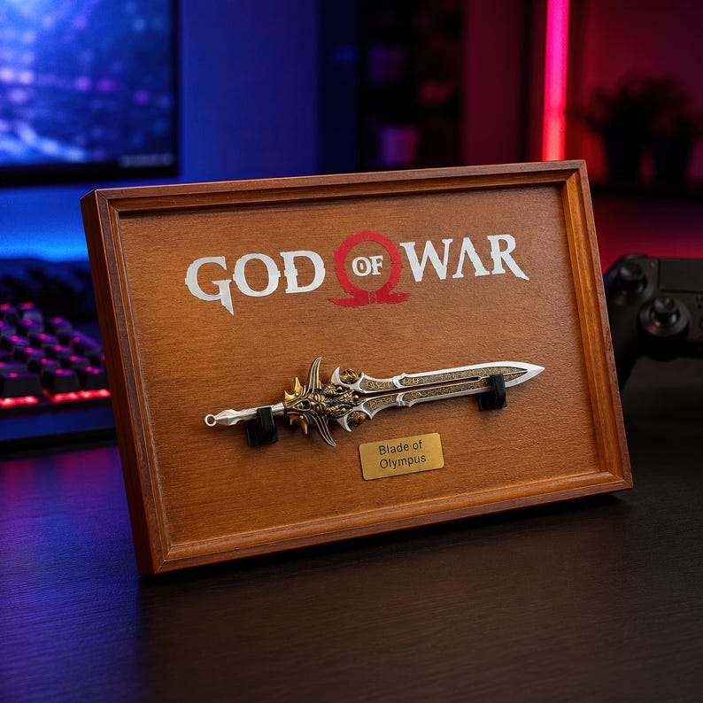 May include: A framed wooden display featuring the words "GOD OF WAR" and a replica of the "Blade of Olympus" sword. The sword is silver and gold, mounted on a wooden plaque. A small gold plaque reads "Blade of Olympus".