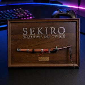 Può includere: Un espositore in legno incorniciato con il titolo del gioco "SEKIRO: SHADOWS DIE TWICE" in lettere argentate. Sotto, una replica di katana montata, con manico marrone, finiture nere e impugnatura avvolta in rosso. Una piccola targa con testo giapponese sotto.