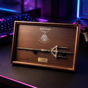 May include: A framed display featuring a detailed replica of Mohgwyn's Sacred Spear from Elden Ring. The spear is metallic with intricate detailing, mounted on a dark wooden background. The frame includes the game's logo and the spear's name on a small plaque.