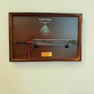 Op de afbeelding: Een ingelijste presentatie van een zwaard uit de videogame Elden Ring. Het zwaard is gemonteerd op een donkere houten achtergrond, met het logo van het spel erboven. Een plaquette leest "Marais Executioner's Sword". Een rode ketting hangt aan het zwaard.