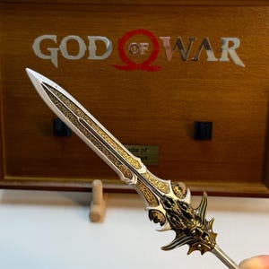 May include: A detailed, decorative sword with a silver blade and gold accents. The hilt features intricate designs and a dragon-like motif. The background displays the words "GOD OF WAR" in silver letters on a wooden surface.