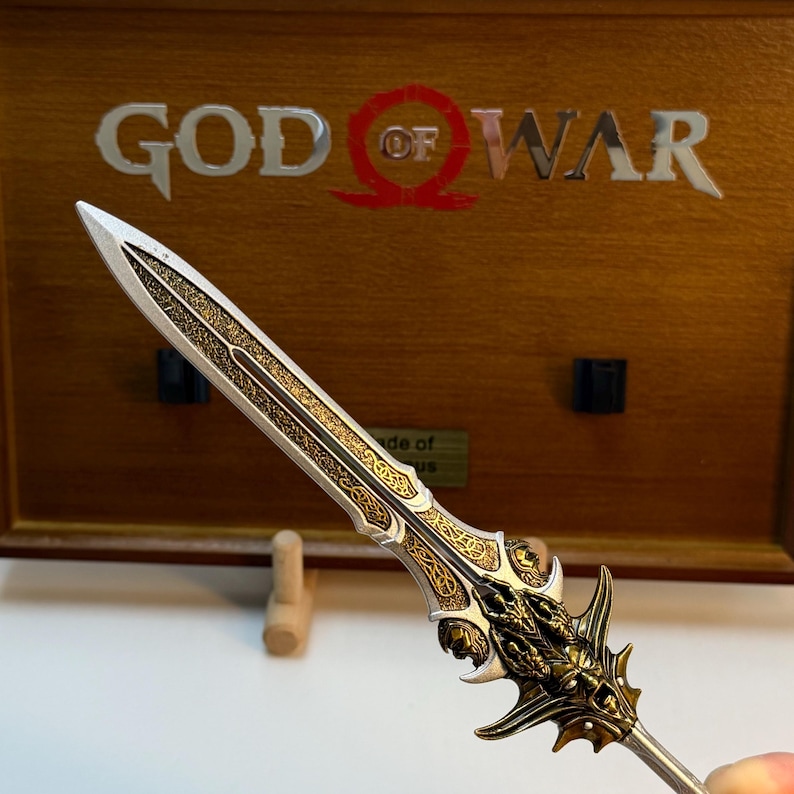 May include: A detailed, decorative sword with a silver blade and gold accents. The hilt features intricate designs and a dragon-like motif. The background displays the words "GOD OF WAR" in silver letters on a wooden surface.