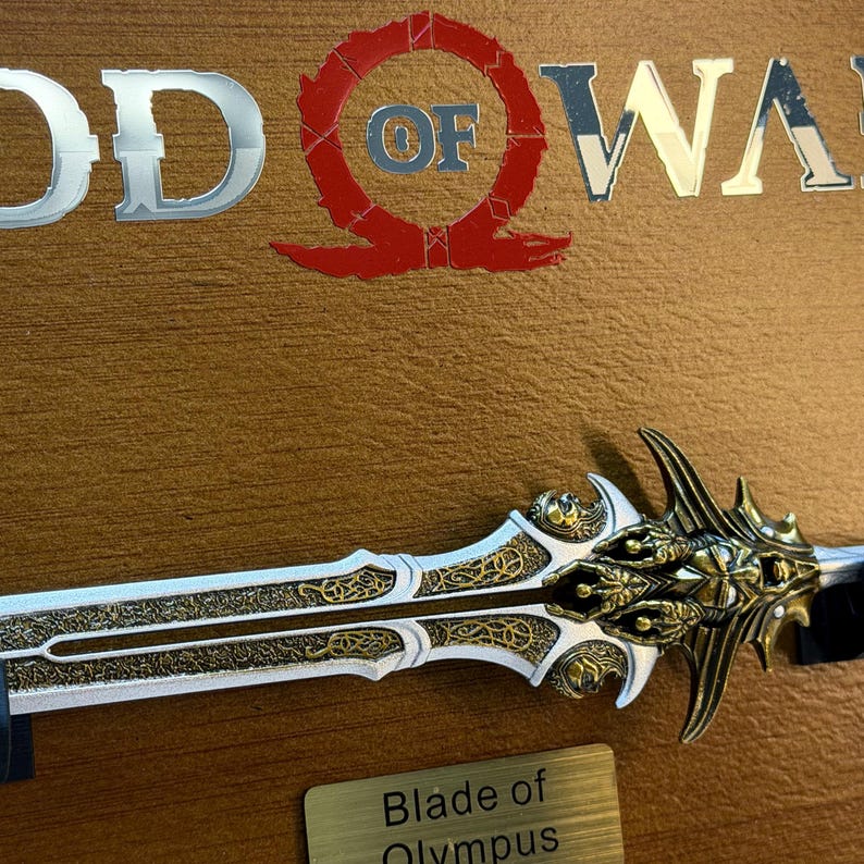 May include: A detailed image of the Blade of Olympus, a prop from the God of War video game. The sword features a gold and silver design with intricate patterns. The God of War logo is visible in the background.