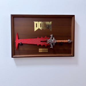 Op de afbeelding: Een wanddisplay met een rood Crucible Blade zwaard met gegraveerde markeringen, een metalen gevest en een beige handvat. Het houten frame toont een gouden DOOM-logo aan de bovenkant en een plaatje met Crucible Blade in het midden onderaan.