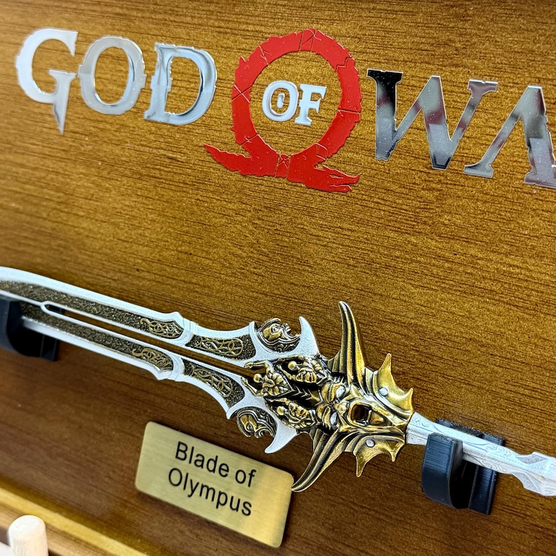 May include: A decorative sword, the Blade of Olympus, with a gold and silver design. The sword is displayed on a wooden surface with the words "GOD OF WAR" in silver letters and a red emblem.