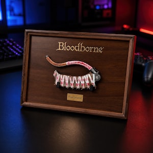 May include: A framed display featuring a miniature Saw Cleaver from Bloodborne mounted on a dark wood board. The cleaver has a metallic blade with red splatter patterns and a curved handle. The text Bloodborne is printed in gold above, with a Saw Cleaver label below.
