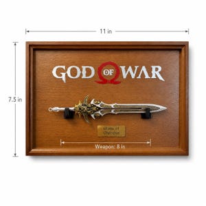 May include: A framed wooden display featuring the "God of War" logo and a replica of the "Blade of Olympus." The sword is 8 inches long, with intricate gold and silver detailing. The frame is 11 inches wide and 7.5 inches tall.