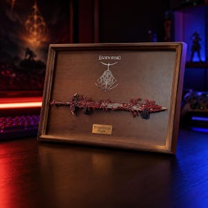 Op de afbeelding: Een ingelijst kunstwerk met een gedetailleerde replica van het Blasphemous Blade uit Elden Ring. Het zwaard heeft rode en zilveren details en is gemonteerd op een houten achtergrond. Het Elden Ring-logo en de tekst "Blasphemous Blade" zijn ook zichtbaar.