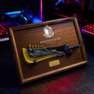 Puede incluir: Un marco de madera con el logotipo de Monster Hunter World y una réplica de la Great Wyvern Jawblade. La espada tiene una hoja metálica oscura texturizada con bordes amarillos translúcidos y un mango negro. Una placa dorada dice Great Wyvern Jawblade.