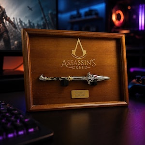 May include: A wooden display frame featuring the Assassin's Creed logo engraved at the top. A miniature silver-toned Spear of Leonidas replica is mounted horizontally in the centre, with a small plaque below reading Spear of Leonidas.