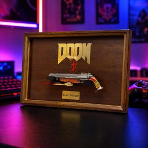 May include: A wooden display frame featuring a metallic Super Shotgun replica mounted on a dark wood panel. The gold DOOM logo is printed above the shotgun, and a small gold plaque below reads Super Shotgun. The frame has a natural wood finish.