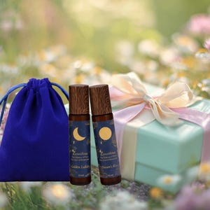 May include: A display of two essential oil roll-on bottles with wooden lids, a blue velvet drawstring bag, and a pale blue gift box tied with a pink ribbon. The bottles feature a moon and star design. The background is a blurred floral scene.