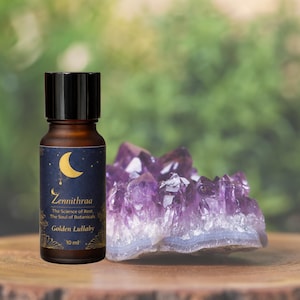 Puede incluir: Una botella de aceite esencial marrón con tapa negra y etiqueta con luna creciente y estrellas. La etiqueta dice "Zennithraa Golden Lullaby 10 ml." Un gran cristal de amatista púrpura está junto a la botella.