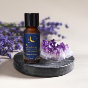 Może przedstawiać: Brązowa butelka olejku eterycznego z czarną nakrętką i etykietą z księżycem i gwiazdami. Butelka stoi na ciemnoszarej kamiennej podstawie z fioletowym kryształem ametystu i gałązkami lawendy. Etykieta głosi "Moon Whisper".