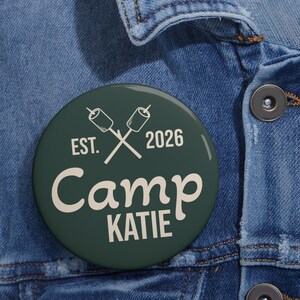 May include: A circular dark green pinback button featuring a cream-colored graphic of two crossed marshmallows on sticks. The text reads EST. 2026, Camp, and KATIE in a mix of serif and script fonts. The button is attached to a blue denim fabric.