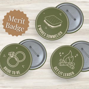 Op de afbeelding: Een verzameling olijfgroene speldjes met zilveren pin backs. De speldjes tonen witte lijntekeningen van een s'more, trouwringen en een kampvuur, met de tekst "S'mores Sommelier", "Bride to Be" en "Get Lit Leader". Een bruine Merit Badge is ook afgebeeld.