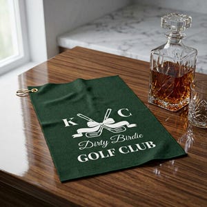 May include: A dark green golf towel with white text reading "KC Dirty Birdie GOLF CLUB". The towel has a gold-coloured hook and a graphic of crossed golf clubs. A crystal decanter and glass are in the background.