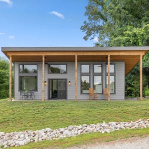 May include: A modern, single-story house with a gray exterior, large windows, and a wooden porch roof. The house is surrounded by green grass and trees, with a blue sky overhead. Two chairs are visible on the porch.