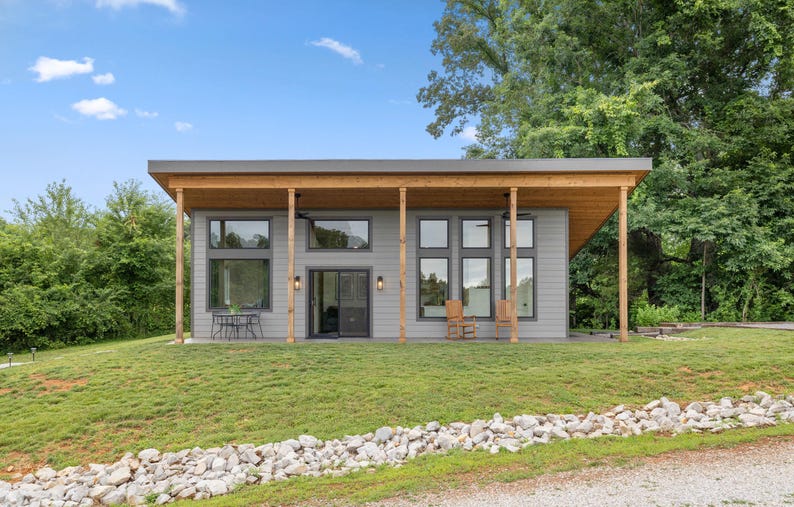 May include: A modern, single-story house with a gray exterior, large windows, and a wooden porch roof. The house is surrounded by green grass and trees, with a blue sky overhead. Two chairs are visible on the porch.