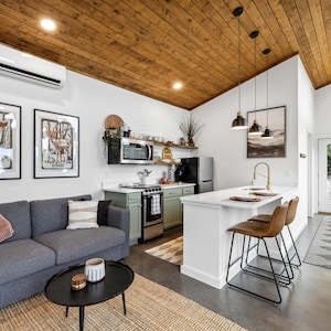 May include: Interior shot of a modern living space with a gray sofa, black coffee table, and a kitchen area. The kitchen features a white countertop, bar stools, and a small refrigerator. Artwork hangs on the white walls, and the ceiling is wood-paneled.