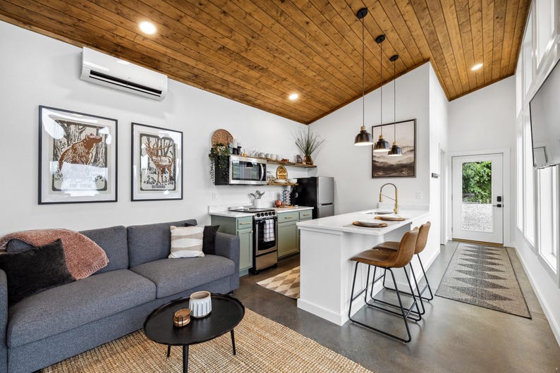 May include: Interior shot of a modern living space with a gray sofa, black coffee table, and a kitchen area. The kitchen features a white countertop, bar stools, and a small refrigerator. Artwork hangs on the white walls, and the ceiling is wood-paneled.