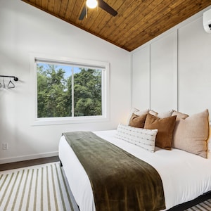 May include: A bedroom with a white bed, a green blanket, and several brown and beige pillows. The room has a wooden ceiling, a window with a view of trees, and a light-colored dresser. A striped rug is on the floor.