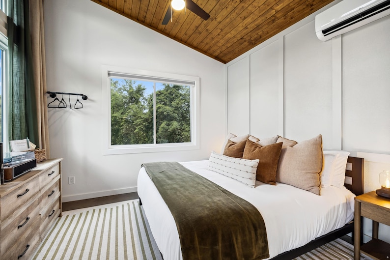 May include: A bedroom with a white bed, a green blanket, and several brown and beige pillows. The room has a wooden ceiling, a window with a view of trees, and a light-colored dresser. A striped rug is on the floor.