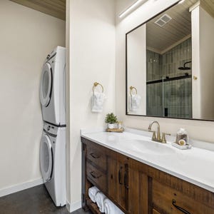 May include: A modern bathroom with a white countertop, a wooden vanity, and a large mirror. A stacked washer and dryer unit is visible on the left. The bathroom has gold fixtures and a small decorative plant.