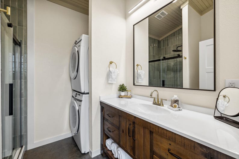 May include: A modern bathroom with a white countertop, a wooden vanity, and a large mirror. A stacked washer and dryer unit is visible on the left. The bathroom has gold fixtures and a small decorative plant.