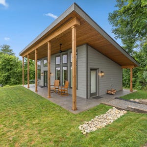 May include: A modern, single-story house with a gray exterior, wooden porch supports, and a sloped roof. The house has large windows and a covered porch with wooden rocking chairs. The surrounding area features a green lawn and a stone pathway.