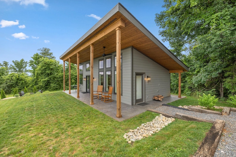 May include: A modern, single-story house with a gray exterior, wooden porch supports, and a sloped roof. The house has large windows and a covered porch with wooden rocking chairs. The surrounding area features a green lawn and a stone pathway.