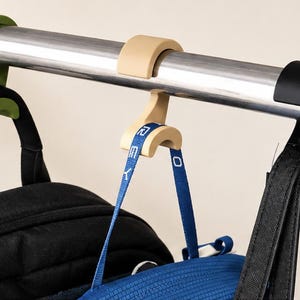 May include: Three C-shaped plastic hooks in olive green, beige, and black are mounted on a horizontal silver metal rod. The beige hook supports a blue strap with white text that reads "EMEY". Black bags hang from the green and black hooks.