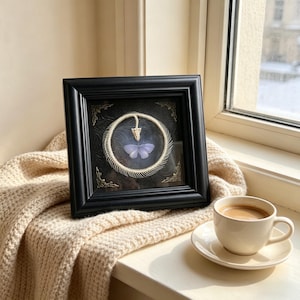 May include: A framed artwork featuring a light purple butterfly within a circular bone structure, set against a dark background. The black frame has ornate corner details. A cup of coffee sits on a saucer nearby, resting on a beige blanket.