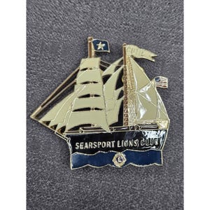 Pode incluir: Um broche de metal dourado em forma de veleiro com velas cor de creme. O casco é azul escuro com o texto SEARSPORT LIONS CLUB em branco. Uma pequena bandeira no topo diz CUTTY e uma pequena bandeira americana é visível à direita.