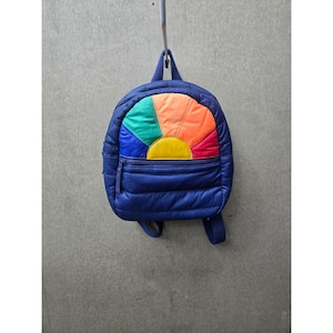 May include: A small, quilted navy blue backpack featuring a vibrant, multi-colored sunburst design on the front pocket. The sunburst pattern consists of segments in bright green, orange, red, and yellow. It has a top handle and adjustable straps.