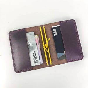 Peut inclure: Un portefeuille en cuir violet foncé doté de deux poches latérales et d'un système de cordon élastique jaune central. La poche gauche contient une carte avec le texte American Express, tandis que la droite contient une carte noire avec un logo blanc.