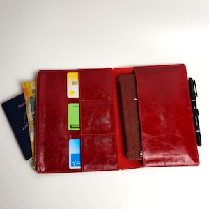 May include: A red leather travel organizer featuring multiple interior card slots, a zippered compartment, a vertical sleeve holding a dark blue passport, and a side pen loop with a black pen. The interior includes a brown notebook.