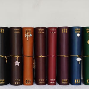 Könnte beinhalten: Eine Sammlung von Lederjournalen in verschiedenen Farben, darunter Schwarz, Braun und Grün. Jedes Journal ist zylindrisch mit goldenen Akzenten und dekorativen Anhängern. Die Journale sind etwa 20 cm hoch.