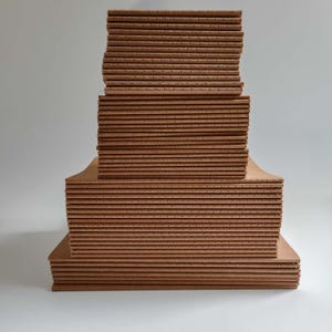 Puede incluir: Una pila de cuadernos de papel marrón claro dispuestos en tres niveles distintos de anchos variables. Cada cuaderno presenta un lomo cosido visible con perforaciones espaciadas uniformemente a lo largo del borde, creando una textura constante.