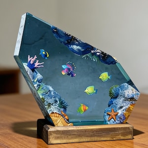 May include: A decorative resin sculpture featuring a miniature underwater scene. The artwork showcases colorful fish, coral, and a starfish within a clear, angled block. The base is a rectangular wooden stand. The overall color palette includes blues, greens, and yellows.